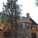 Exterior of handcrafted log home with large covered entry framed with log truss, octagon sunroom and stone chimney set on stone faced basement on hillside in California