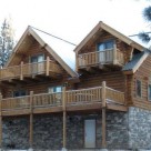 Exterior side view of handcrafted log home with two dormers with balconies set above large deck with log rails on stone covered lower level.