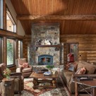 Log home greatroom with stone fireplace beyond cozy seating area. Log staircase leading to open loft with log railings and bear carvings in log posts.