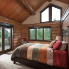 Master bedroom with king size bed in handcrafted log home with trapezoid windows in gable end.