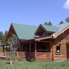 Exterior photo of handcrafted chink style log home with green metal roof, covered porches dormer and balcony with log railing.