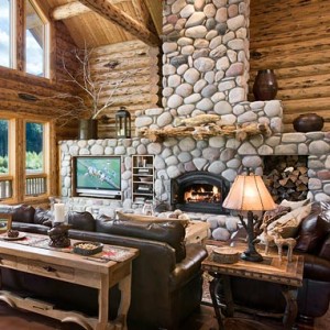 The interiors of a log cabin great room, complete with a cozy rock fireplaces and beautiful log beams.