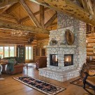 3 sided fireplace in handcrafted log home with wood floors, rocking chair and cathedral ceiling supported by massive log trusses.