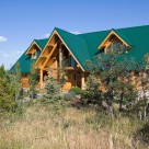 Handcrafted log home with green metal roof with small dormers on each side of center roof supported by log post and beams.