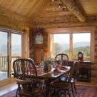 Beautiful dining table and chairs set on area rug in handcrafted log home with wood floors and large windows with views to forest beyond.