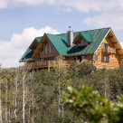 Exterior photo of handcrafted log home with green metal roof, steep pitched gables and small doghouse dormers on either side of main roof line.
