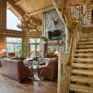 Great room of luxury log home with curved log staircase, massive stone fireplace and massive bay windows surounded by large log columns.