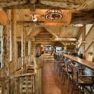 Open loft in luxury log home with long bar and barstools and log posts supporting log beams in ceiling.