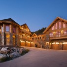 Stunning twilight photo of luxury log home and log guesthouse set above 5 car stone garage. Octagon sunroom set on stone pillars to side of driveway.