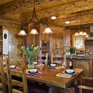 Heavy wood dining table and chairs with chandelier above and luxury log home kitchen in background with exposed logs and pine board ceiling.