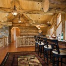 Fancy bar with barstools in open loft of log home with massive logs, pine ceiling over exposed log beams, hardwood floors with area rug.