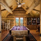Custom made pool table in loft of handcrafted log home viewed through open glass doors with full log gable and exposed roof beams.