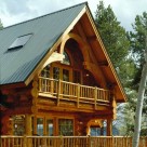 Close up exterior photo of log home balcony framed in timber hammerbeam truss and log rails with french doors in gable behind.