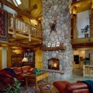 Great room with soaring cathedral ceilings with red leather sofa and log endtables set on area rug in front of massive stone fireplace with antique snowshoes hung above log mantle.