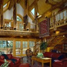 Custom log home greatroom with cathedral ceilings supported by character log posts and log purlins. Open loft above with log railings and archtop window in gable. View to Colorado forest through frenchdoors at end wall of log home with log staircase to loft on right.