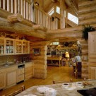 Interior of handcrafted log home with custom cabinets and view through log archway to stone fireplace in greatroom with transom windows.