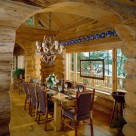 Massive log slab dining table with rustic log chairs and antler chandelier set in handcrafted log home dining room viewed through log archway