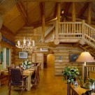Interior of handcrafted log home with massive log plank dining table, antler chandelier, log stairs to open loft with log railings and cathedral ceilings supported by log posts and purlins.