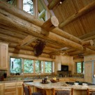 Custom log home kitchen with custom cabinets, tile counters, breakfast bar with log barstools. Exposed log beams above with glass in gable and elk head mount on wall.