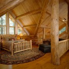 Log bed set on oval area rug in open loft of custom log home with log post and beam ceiling and white gable with trapezoid windows.
