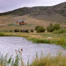 Custom log home on hillside viewed across pond with cattails and willows.