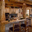 Custom log home kitchen with pine cabinets viewed though log posts on each end of breakfast bar with thin set stone face and live edge log slab counter.