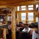 Handcrafted log home greatroom with log mantle on fireplace and large windows trimmed in log post and beam.