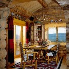 Log home dining room with antler chandelier above custom wood table set on hardwood floors viewed through archway in log wall