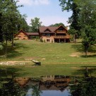 Beautiful log home with log garage attached by breezway viewed from across pond with canoe on shore and lush green lawn leading to home.