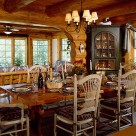 Large wooden dining table with high back white wooden chairs in dining room of log home with archway leading to sunroom with large grilled windows and green corner china hutch.
