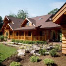Exterior of custom log home with covered porch leading to breezway attaching log garage. Fresh green landscaping accents walkway.