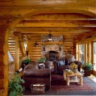 Sitting area with leather sofa's on area rug and wood stove set in stone chimney viewed through log archway in custom log home.