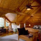 Loft floor bedroom in handcrafted log home with gable dormer streaming light onto wicker rockers, cozy bed and wood bench at foot of bed.