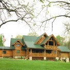 Exterior photo of handcrafted log home with green metal roof with multiple roof lines, covered balcony and log railings.