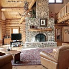 Log home greatroom with massive stone fireplace with log mantle, large area rug and three comfy chairs. Log beams supporting loft above with log raiings