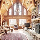 Log home greatroom with large dry stack masonry fireplace with log mantle, large red patterned area rug with sofa, large windows in end wall with trapezoid windows above in gable viewing hardwood trees outside.