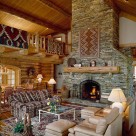 Living room with massive stone fireplace with log mantle in luxury log home with cathedral ceilings, open loft with log railings to left and exposed log purlins with pine ceilings.