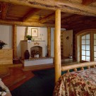 Master bedroom in luxury log home with log bed on green carpet and fireplace set between log posts against white wall. Log beams support pine board ceiling.