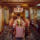 Large glass top dining table set on log frame with upholsted log chairs set on area rug in handcrafted log home with spiral log staircase on right and round top french doors on left.