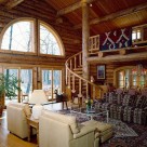 Interior log home living room with log spiral staircase to open loft with log rails, view through french doors and arched windows to Pensylvania forest.