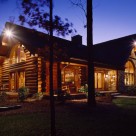 Exterior view of luxury log home at twilight. Home features stone wall at center and handcrafted log construction.