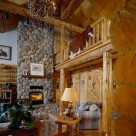 Greatroom with river rock fireplace and hardwood floors in handcrafted log home with open loft above supported by log post and beam and log railings with cowhide rug draped over edge.