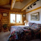 Guest bedroom with sloping pine ceiling in log cabin with track lighting mounted to log beams above.