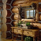 Close up photo of log home interior with massive chinked log wall and corner. Ornate table with drawers is set below large woodframed mirror with elk antlers on either side.