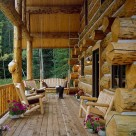 Covered porch at end wall of handcrafted log home. Log posts and beams support cathedral roof above wood benches and flowers in this rustic log home.