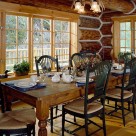 Dining room in handcrafted log home with large wooden table and green highback wood chairs look out through large windows onto deck with log railings and forest beyond.