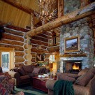 Great room with cozy chairs on green carpet view large riverrock fireplace in Handcrafted log home. Antler chandelier hangs in front of massive log supporting cathedral ceiling..