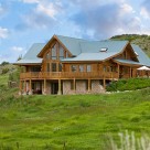 Exterior view of large 3 chamber custom log home with green metal roof set on walkout basement on green sloping land.