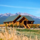 Exterior view of handcrafted log home with wrap around deck, log railings and green roof framed by Colorado Rocky mountains in background.