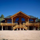 Exterior of custom log home duplex with log truss at covered entry and large decks with log railings. Semicircle windows on either side of center.